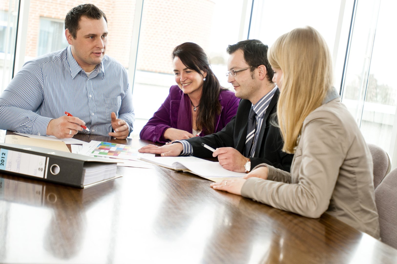 Eine Teambesprechung mit vier Personen (zwei Frauen, zwei Männer). Auf dem Tisch liegt ein Aktenordner.