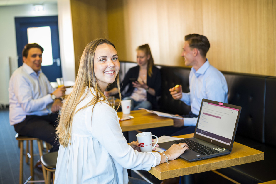 Vier Auszubildende der TRAVE an Bistrotischen. Im Vordergrund lächelt eine junge Frau (Auszubildende) mit Laptop in die Kamera.