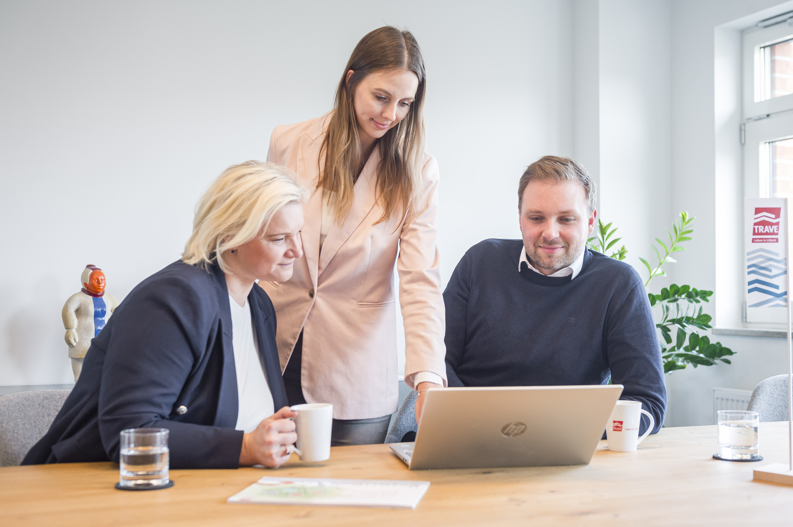 Teamarbeit bei der TRAVE Drei Personen sitzen gemeinsam an einem Tisch und arbeiten an einem Laptop in einem hellen Büro.